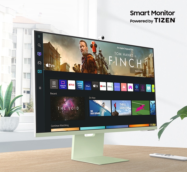 The monitor sits on top of a wooden desk in front of a window surrounded by plants. The words ††Smart Monitor Powered by Tizen'' are shown above the monitor. The screen shows a variety of content apps and individual content choices in a card format. A remote rests next to the monitor.